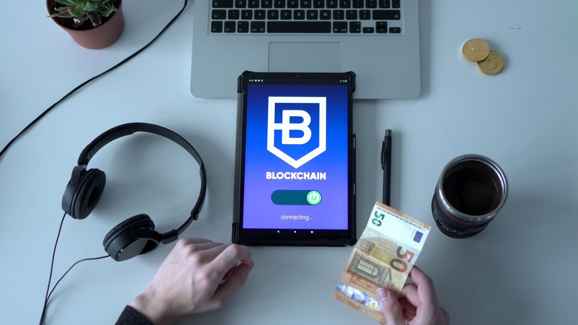 Overhead view of digital tablet with blockchain app, cash, and tech tools on desk.