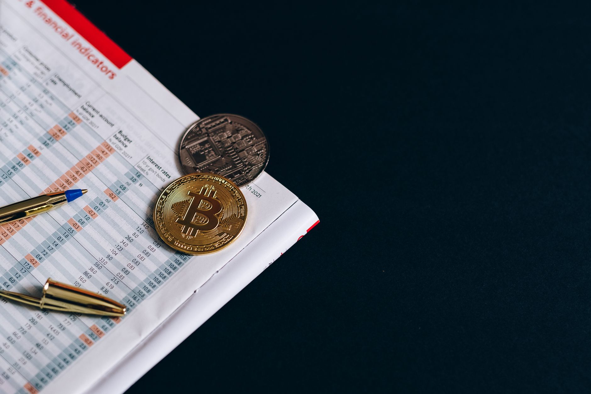 A close-up of Bitcoin with financial indicators and a pen on a black surface.