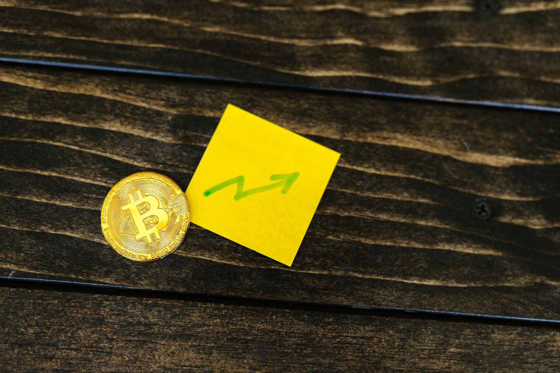 Close-up of a Bitcoin coin with an upward arrow sticky note on a wooden surface.